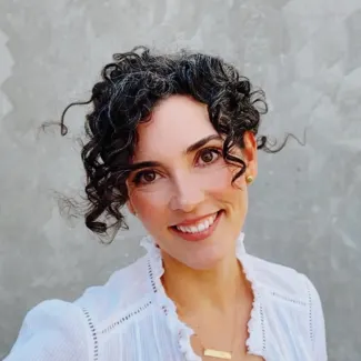 A woman with brunette curly hair wears a white blouse.