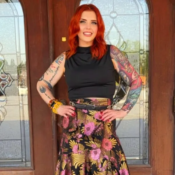 A woman with red hair and her arms akimbo, standing in front of glass doors. 