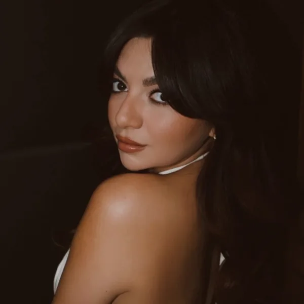 A young woman with long brunette hair looks over her shoulder while wearing a white halter top.