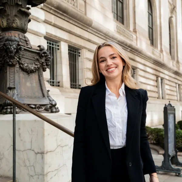 A young woman with blonde hair smiles on the steps of a building.
