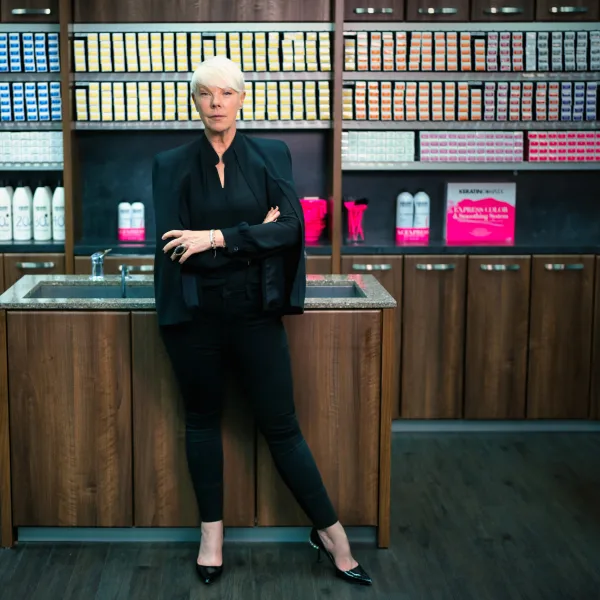 A woman in black and with short blonde hair, with arms crossed, standing in front of shelves of beauty products.