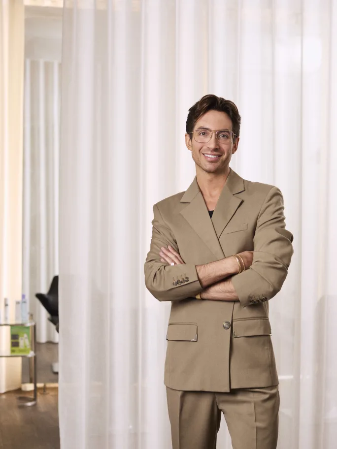 Portrait photo of a smiling man in a tan blazer suit with arms crossed, wearing glasses and gold jewelry, standing in front of sheer white curtains in a salon setting.