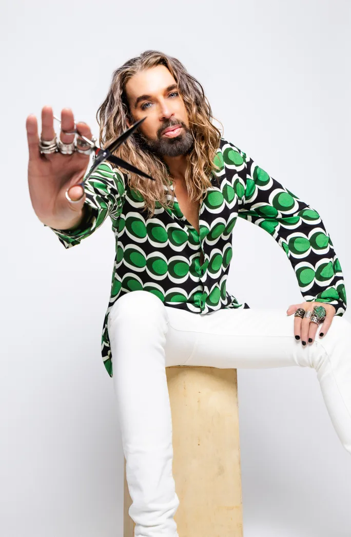A man with long blonde hair, wearing a green shirt and white pants, holds a pair of shears toward the camera.