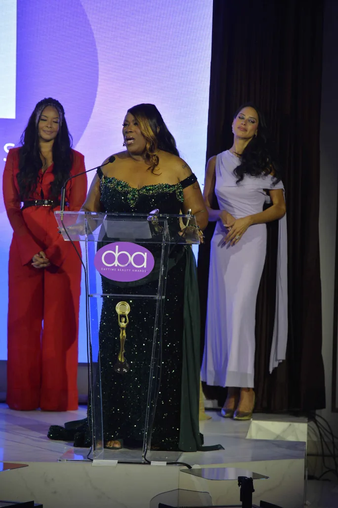 Three women stand onstage as one stands in front of a podium.