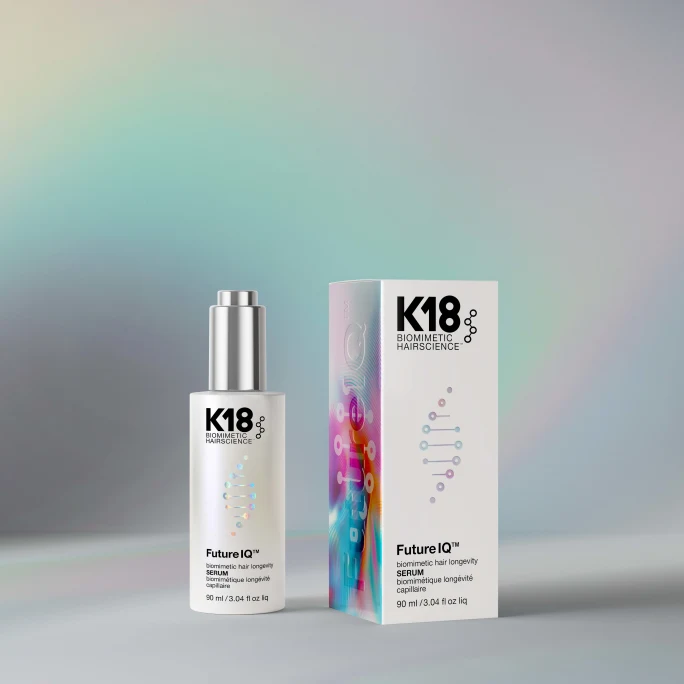 Product photo of a K18 Biomimetic Hairscience Future IQ hair longevity serum bottle (90 ml) standing beside its matching packaging box, both featuring a colorful DNA-strand graphic, set against a soft pastel-rainbow gradient background.