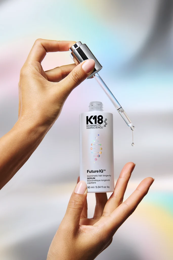 Product photo of a hand holding a K18 Biomimetic Hairscience Future IQ hair longevity serum dropper with a droplet of liquid falling, positioned above the open bottle (90 ml), against a soft pastel-rainbow blurred background.