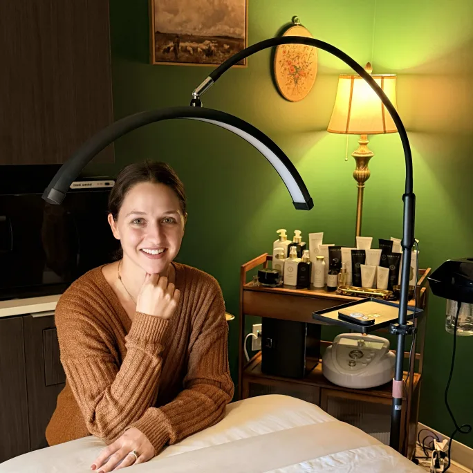 A woman in a brown sweater rests her elbow on a massage table, and smiles.