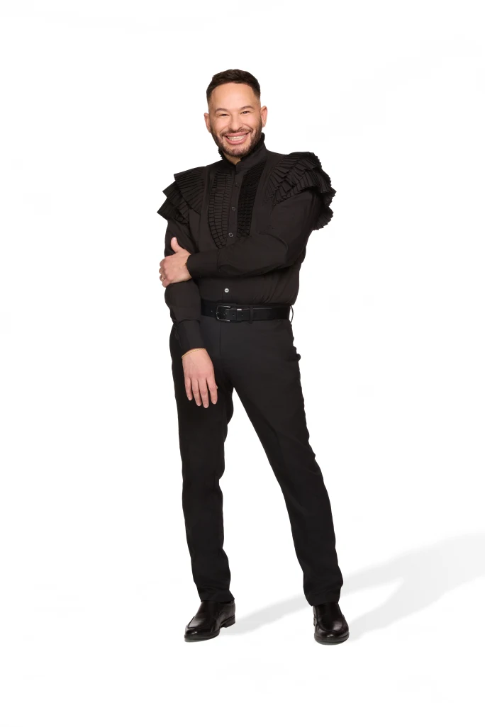 A man stands wearing all black, one arm across his body.