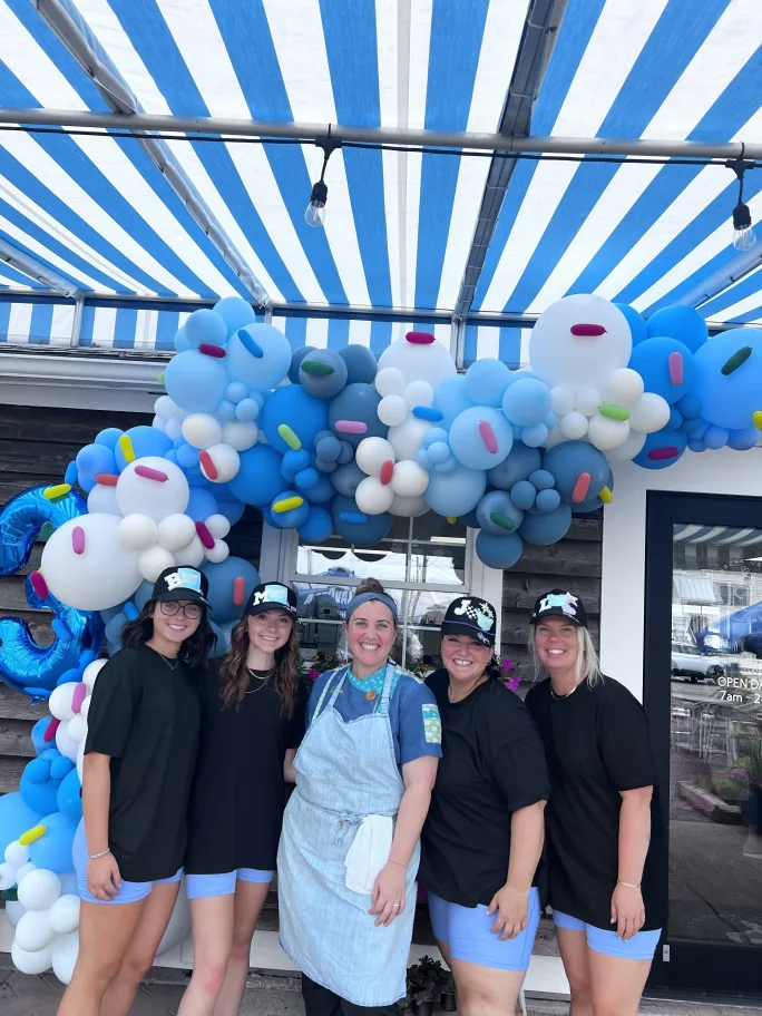 Lillian James team members stand with a baker in front of an arch of blue balloons. 