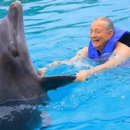 A man holds a dolphin's flippers while in the water. The man is smiling.