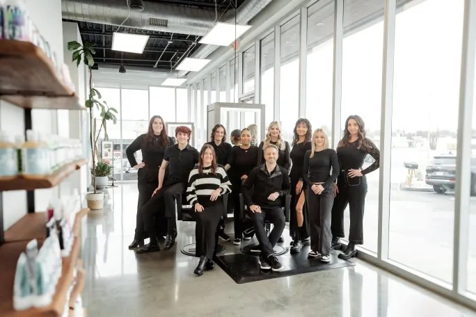 Dressed in black, a group of stylists pose in the salon between retail shelves and big storefront windows.