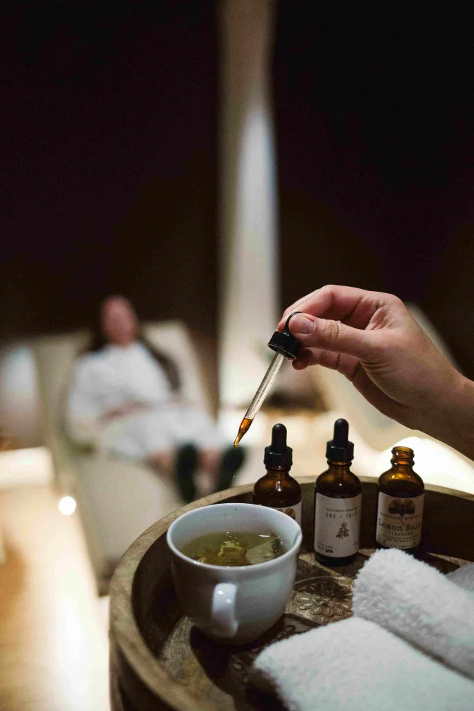 A hostess adds a tincture to a cup of hot tea as a spa guest relaxes in the background.