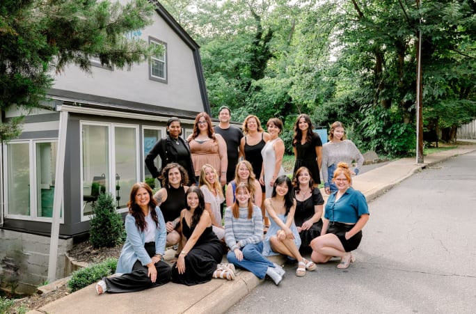 In casual dress, a group of stylists pose outside their salon in the woods.
