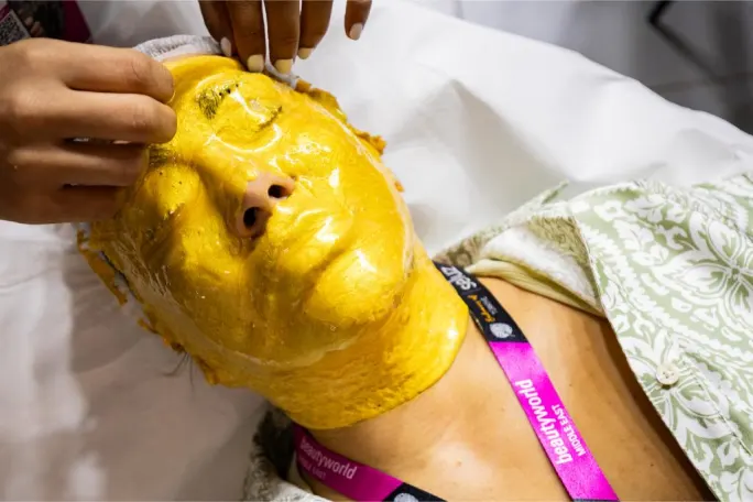 Woman receiving a facial treatment with a gold mask while a practitioner works on her face.