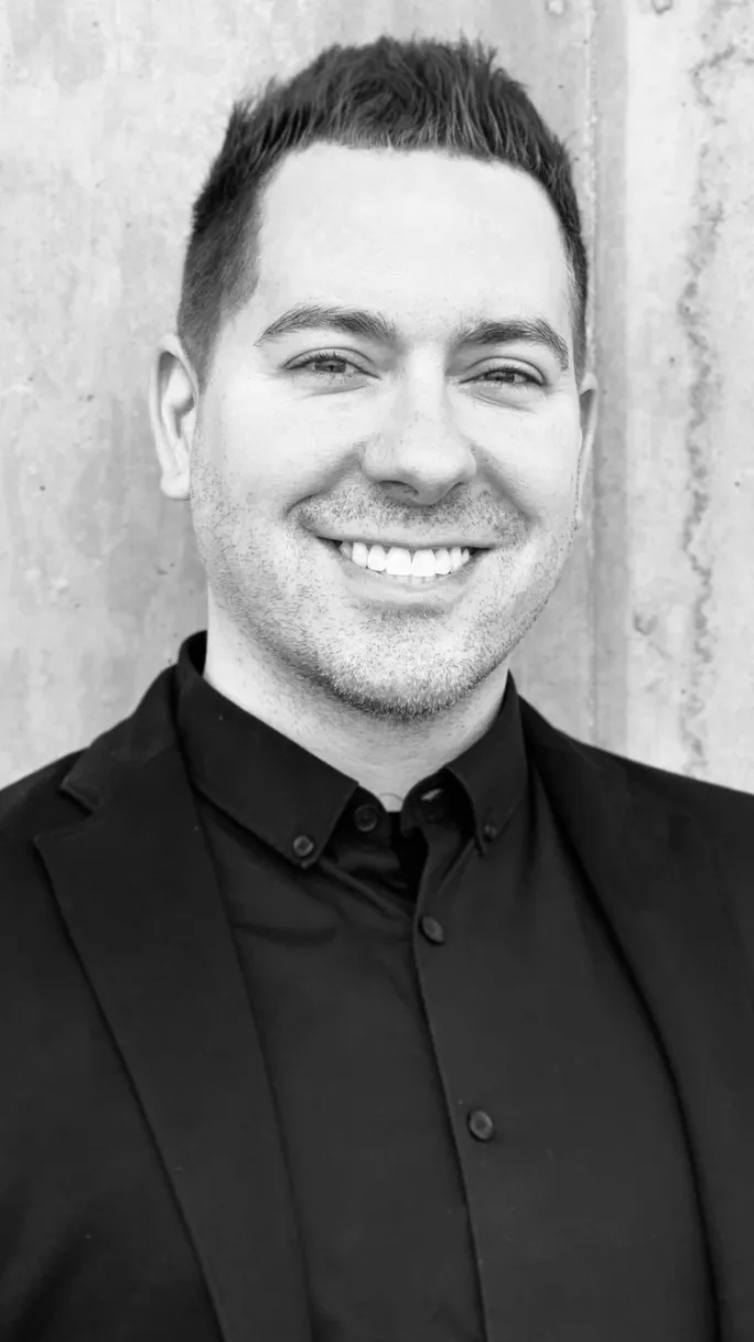 A man in a black suit and shirt, smiling.