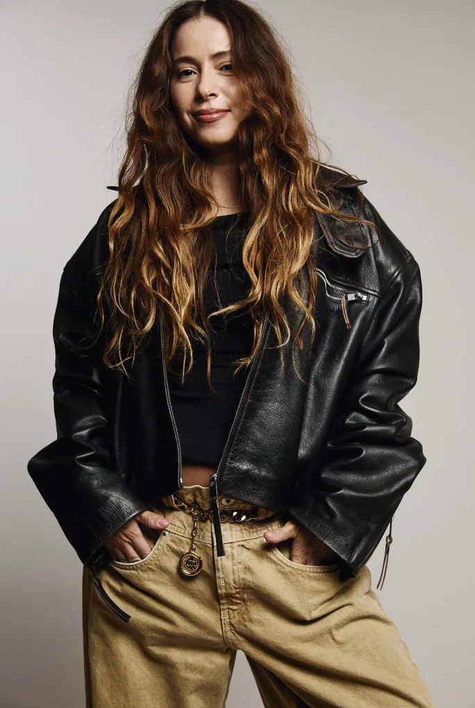 A woman with long blonde hair wearing a black leather jacket and khakis.