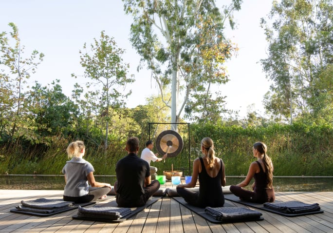 Four people in a circle, meditating, while someone strikes a gong.