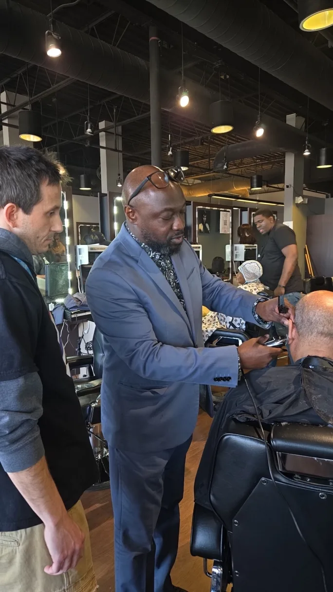 Man teaching a barbering technique to a student in a classroom setting.