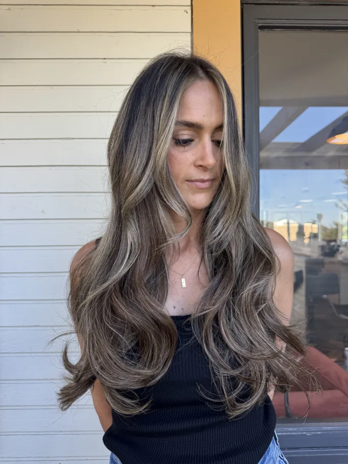 Young woman with long brunette hair enhanced with caramel highlights and soft waves.