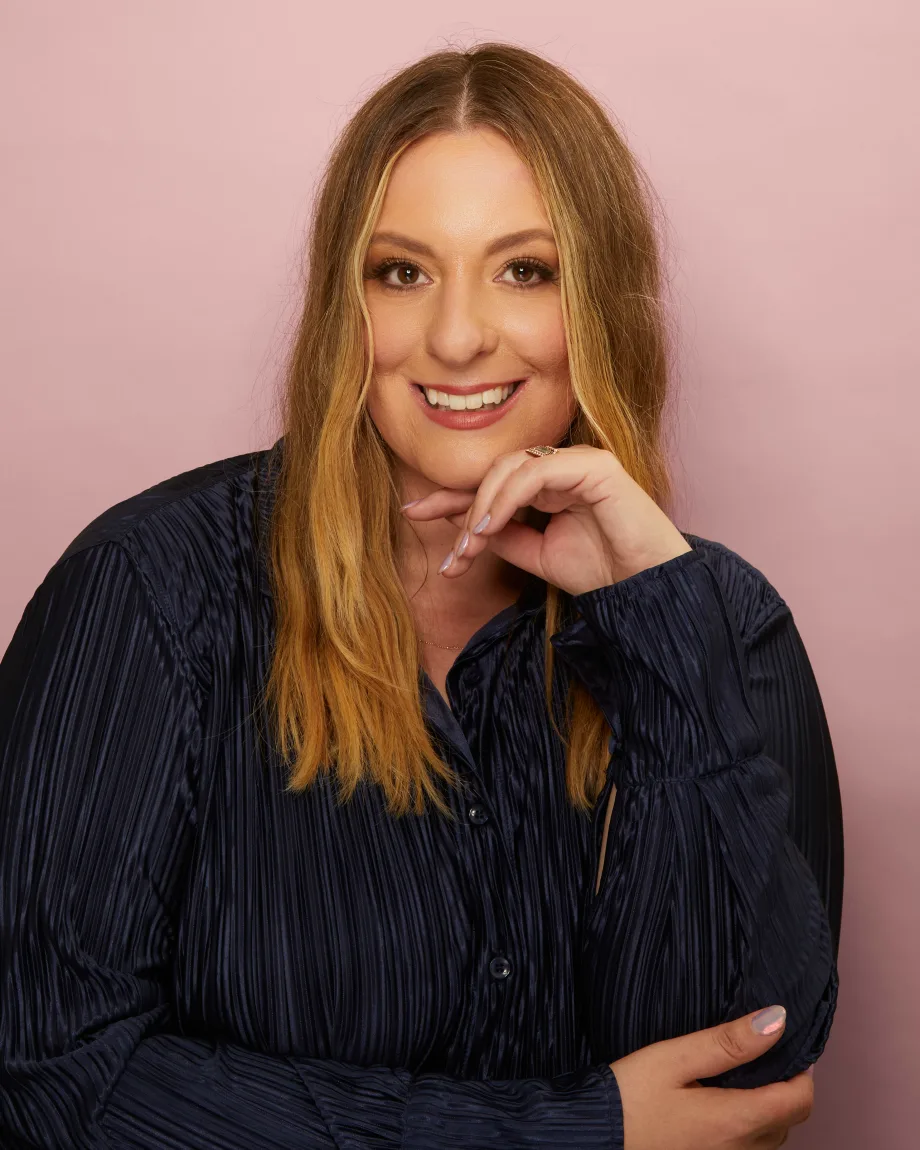 A woman with long blonde hair, wearing a dark blue shirt, smiles while proppng her chin on her hand.