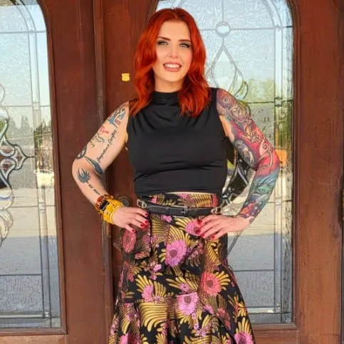 A woman with red hair and her arms akimbo, standing in front of glass doors.