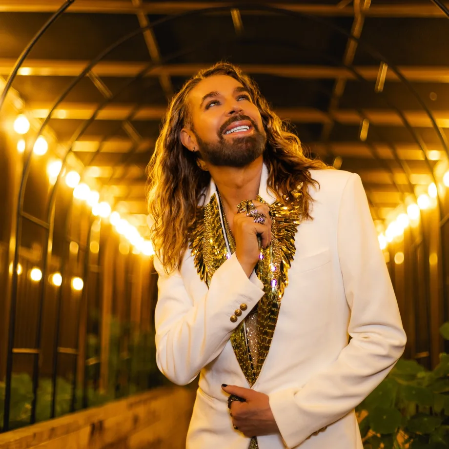 A man with long blonde hair, wearing a white tuxedo jacket, looks upwards.