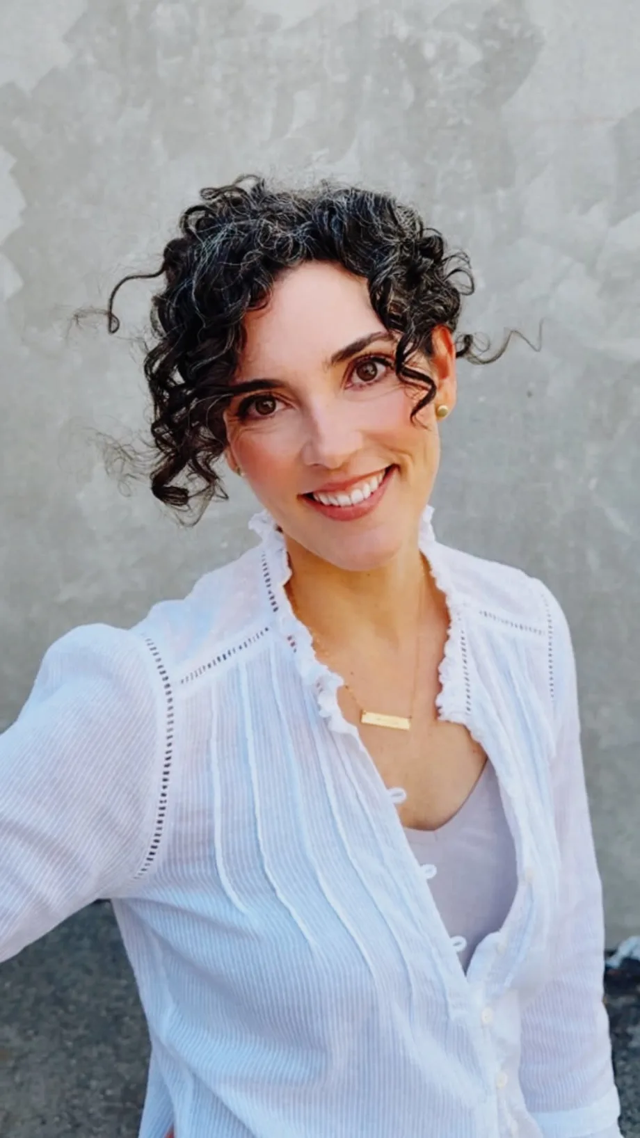 A woman with brunette curly hair wears a white blouse.
