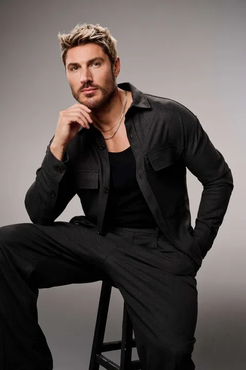 A man wearing black and sitting on a high stool.