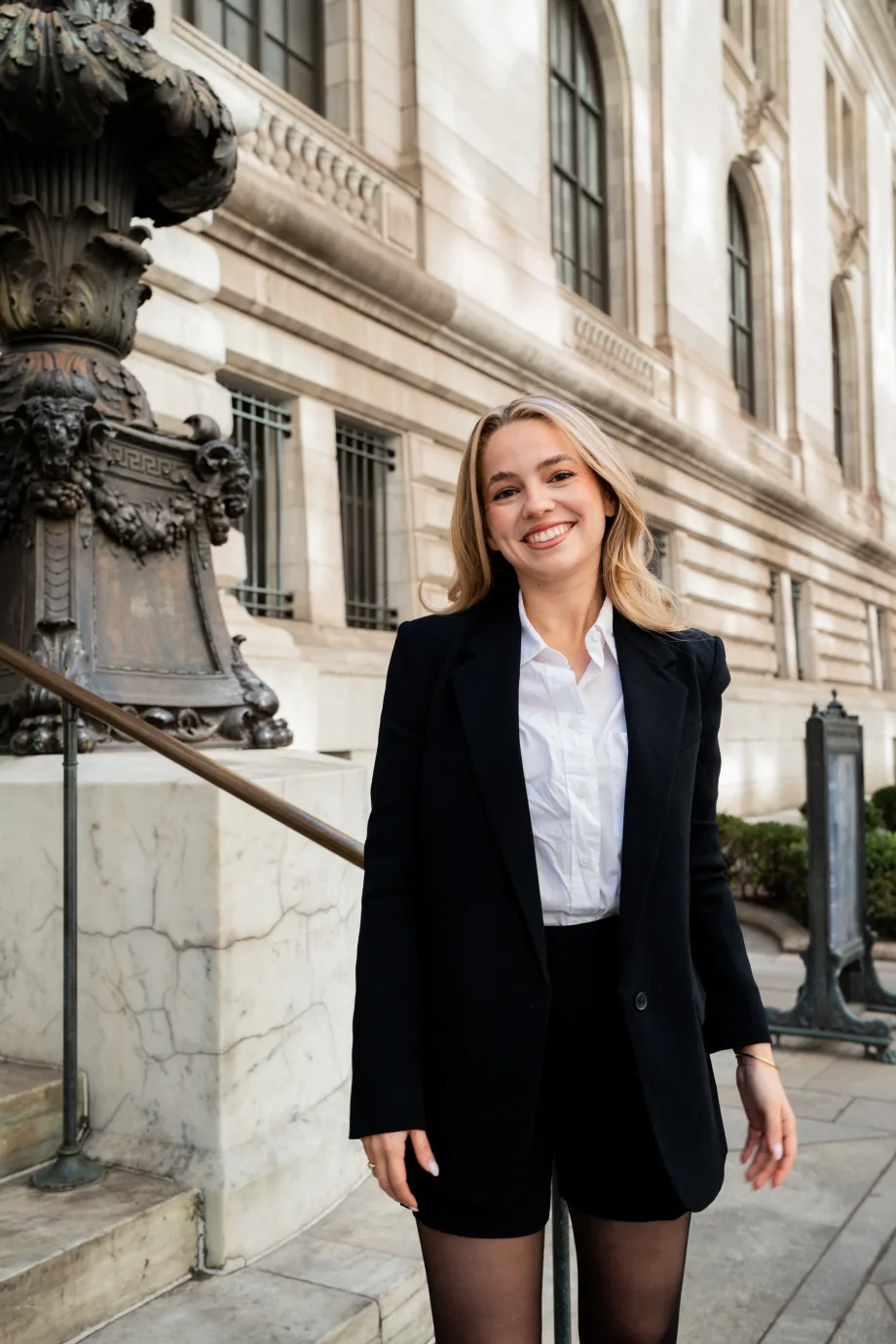 A young woman with blonde hair smiles on the steps of a building.