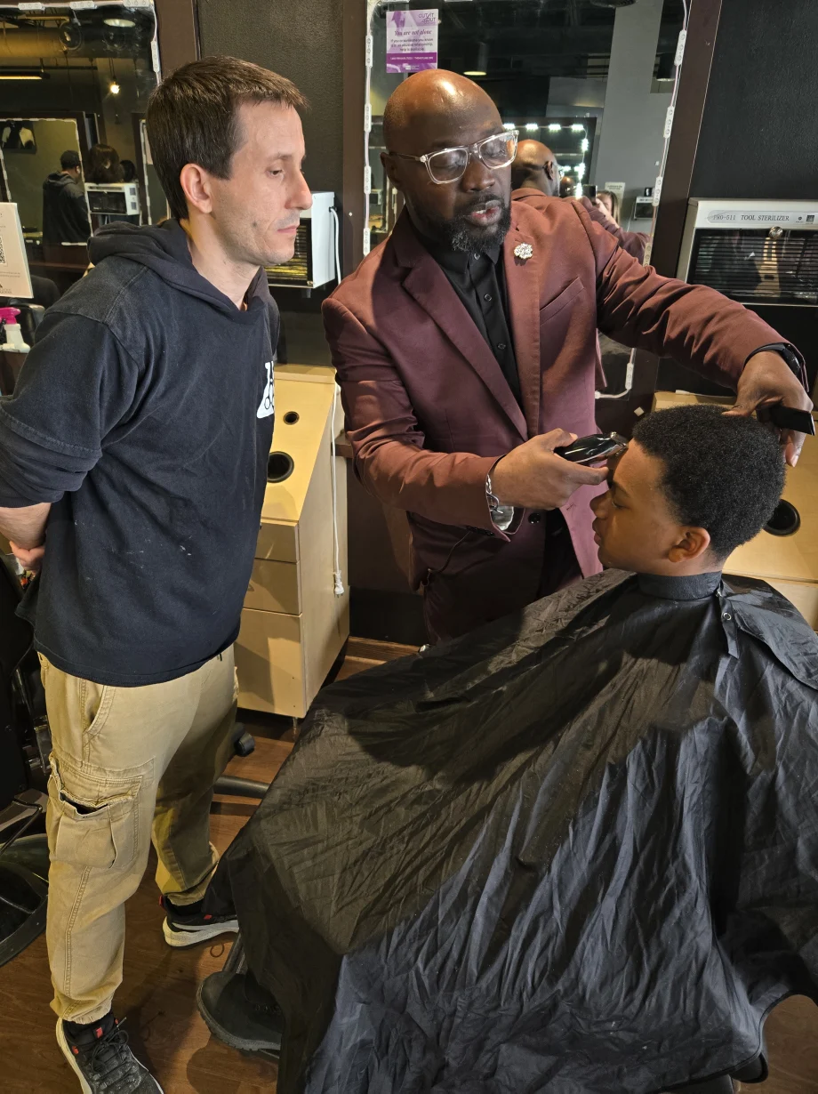 Man teaching a barbering technique to a student in a classroom setting.