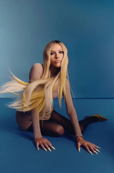 A model with long, blonde hair with yellow accents, sitting on the floor, on set at a photo shoot.