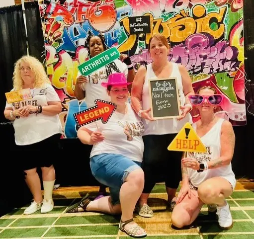 Five nail techs posing together in front of a branded selfie wall at the Nail Tech Event of the Smokies.